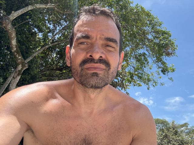 Shirtless XRawForU with a hairy chest, brown beard, and short dark hair, looking slightly confused, under a bright blue sky with scattered clouds and green trees in the background.