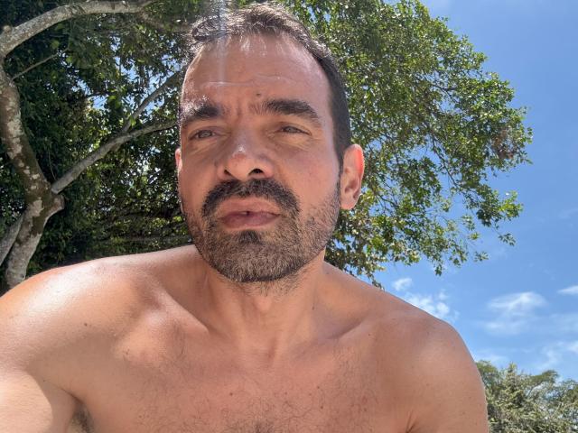 Shirtless XRawForU with a hairy chest and beard, blue eyes, and a slightly confused expression, standing outdoors under a tree with a clear blue sky.