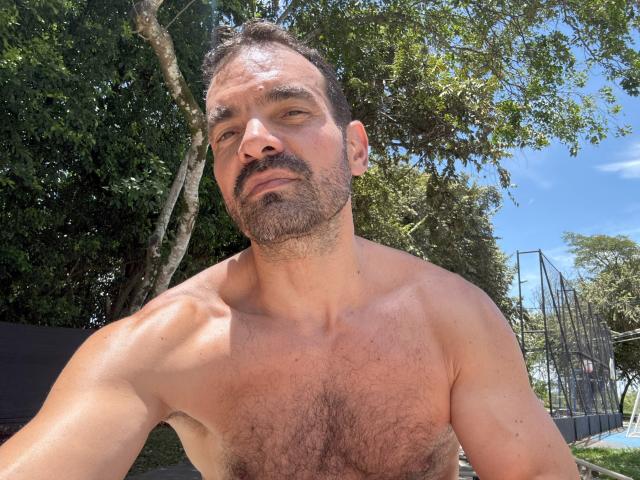 shirtless, hairy, middle-aged, muscular, XRawForU with a beard, short dark hair, looking at camera, outdoors, sunny, blue sky, green trees, metal structure in background.