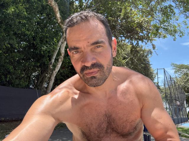 Muscular, shirtless XRawForU with dark hair, beard, and hairy chest, looking at camera with slight smirk, outdoors in sunny, tree-filled area. His ass is partially visible.