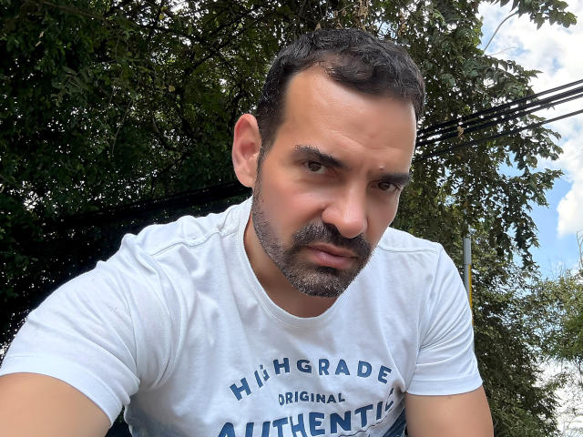 XRawForU has a serious, slightly annoyed expression, dark curly hair, a short beard, and is wearing a white "Hingham Original" t-shirt, looking down at the camera.