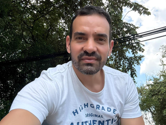 XRawForU with a graying beard, short dark hair, and a white Higrade t-shirt, looking slightly down at the camera, standing outdoors with trees in the background.
