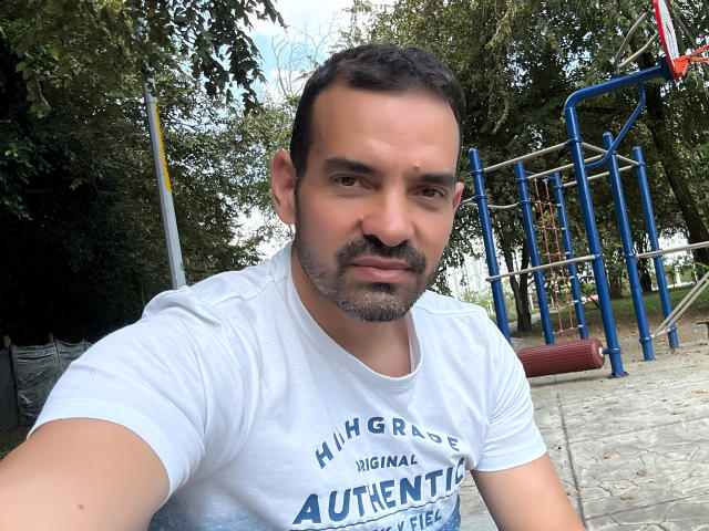 XRawForU has a medium-build, short dark hair, and a beard, wearing a white t-shirt, looking directly at the camera in a park with a playground in the background.
