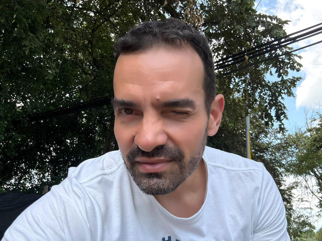 XRawForU has a slightly winking, grumpy face with short dark hair, a beard, and a white shirt. His nose is a little upturned. He's outdoors with trees.