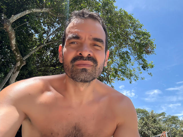 shirtless XRawForU with a slightly tanned, hairy chest, short brown hair, and a beard, looking down with a subtle smile, under a bright blue sky and green trees.