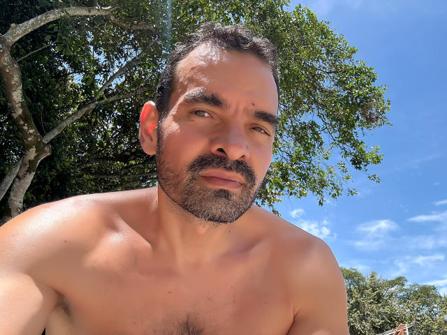 Shirtless XRawForU with a hairy chest and dark beard, blue eyes, and slightly parted lips, standing outdoors with a tree in the background and a bright blue sky.