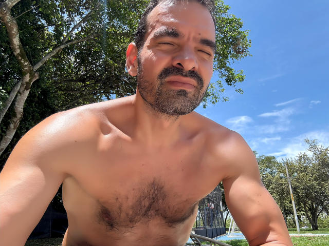 XRawForU, shirtless, muscular, hairy chest, medium brown skin, short dark hair, beard, slightly squinting, standing outdoors, blue sky, trees, sunlight, confident, relaxed, strong jawline, ear piercing.