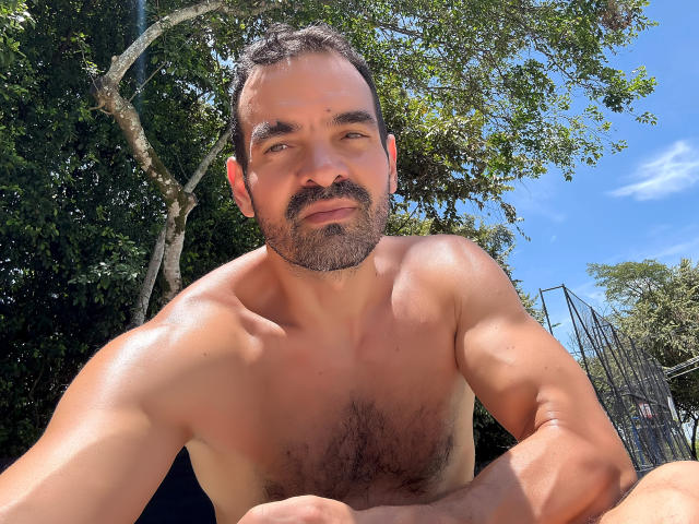 XRawForU, shirtless, muscular, hairy chest, dark hair, beard, slightly tanned, looking down, blue sky, green tree, metal structure, sunny, relaxed, sitting, casual, confident.