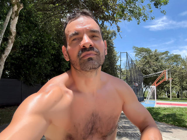 Shirtless XRawForU with a muscular, hairy chest and a slight beard, gazing at the camera, standing outside in a sunny park with a basketball hoop in the background.