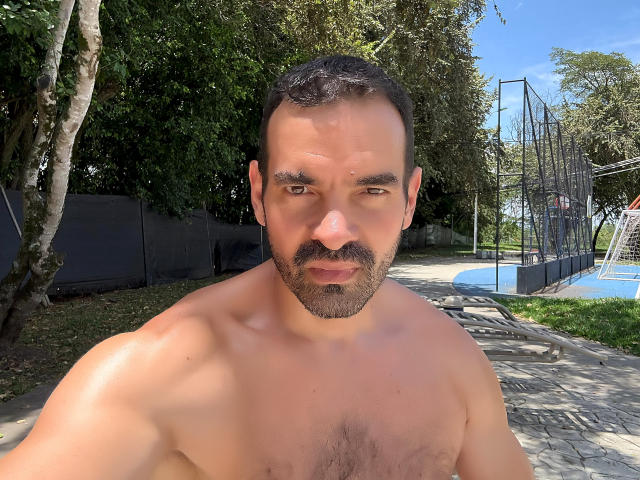 XRawForU has a muscular, shirtless, tan-skinned, bearded, dark-haired guy with a mustache, medium-sized chest hair, and a slight smile. He's taking a selfie outdoors.