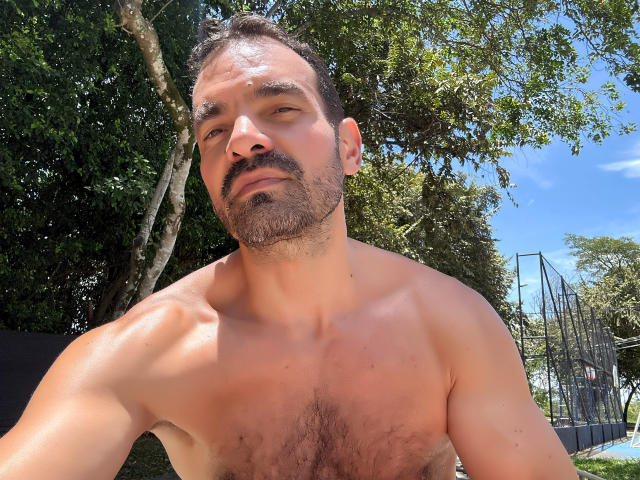 Shirtless XRawForU with a muscular, hairy chest, dark hair, and beard, looking slightly up, outside in sunlight, showing a relaxed, confident expression.