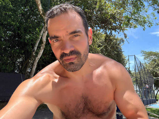 XRawForU has a muscular, hairy chest with a slight tan, short dark hair, and a beard. He's shirtless, looking at the camera with a slight smile.