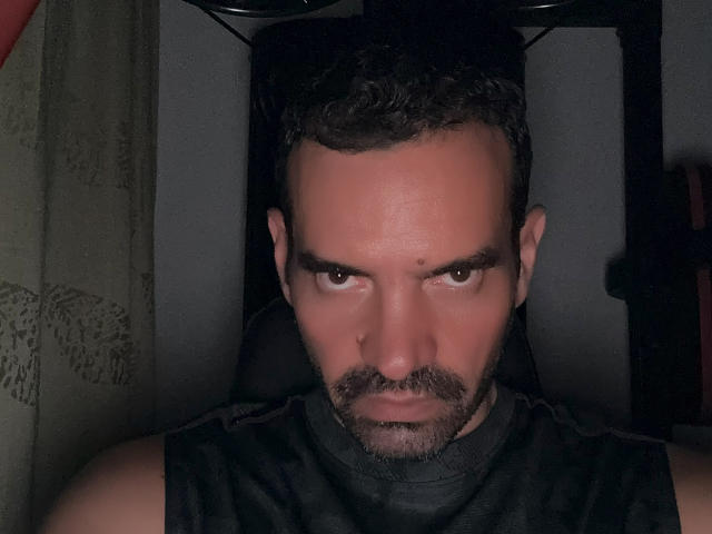XRawForU has a serious, almost angry expression, with dark, curly hair, a beard, and a nose piercing, wearing a black sleeveless top, in a dimly lit setting.