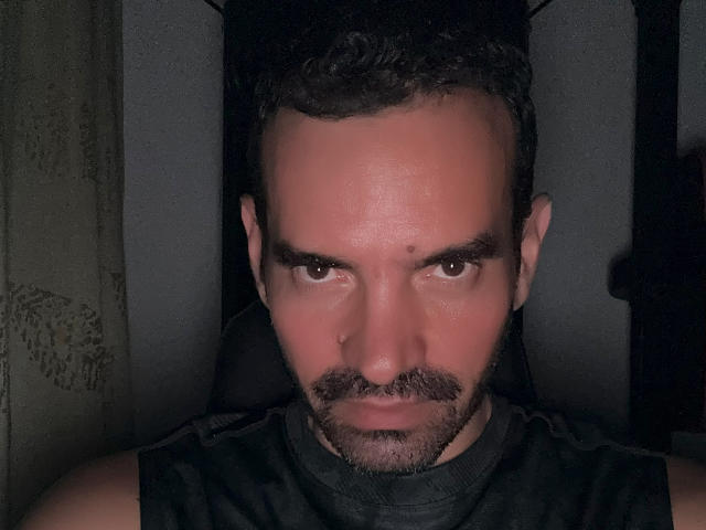 XRawForU has a serious, focused expression with dark, wavy hair and a beard, wearing a black sleeveless shirt, staring intensely at the camera.