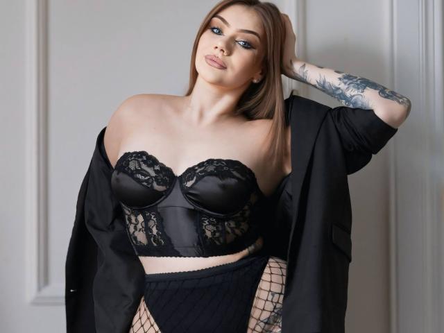 Marrena with tan skin and brown hair, wearing a black lace strapless top, black skirt, and fishnet stockings, black jacket off-shoulder, arm tattoo, pouty lips, confident expression.