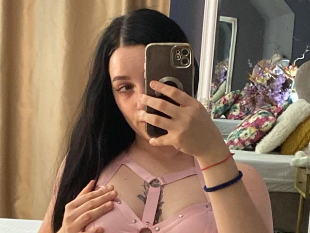 LeahBrooks with long black hair, pink harness top, holding phone in mirror selfie, showing small tattoo under boobs, pink nipples, right hand on chest, blue wristbands.