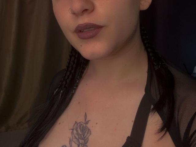 LeahBrooks has braided black hair, dark lipstick, a nose ring, and a rose tattoo on her chest. She's wearing a sheer black top with visible cleavage.