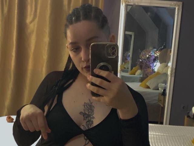 LeahBrooks with braided hair, black top, exposing tits with a rose tattoo, holding a phone, mirror reflection, golden curtain, dark room.