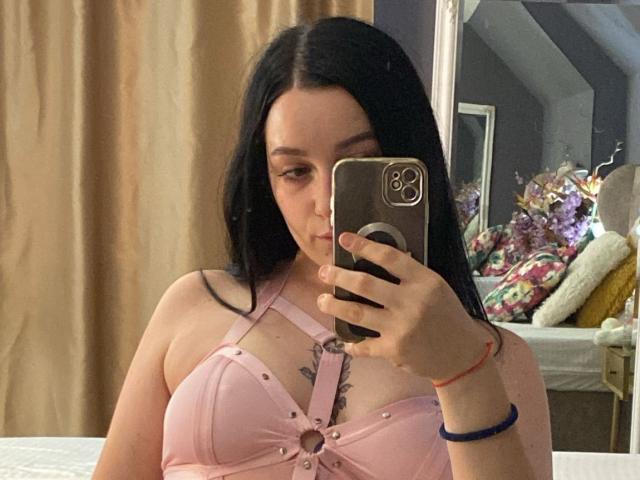 LeahBrooks with black hair, pink strappy bra, holding phone, covering face, medium tits, tattoo below cleavage, pink skin, beige curtain, brown phone case, red bracelet, bed in background.