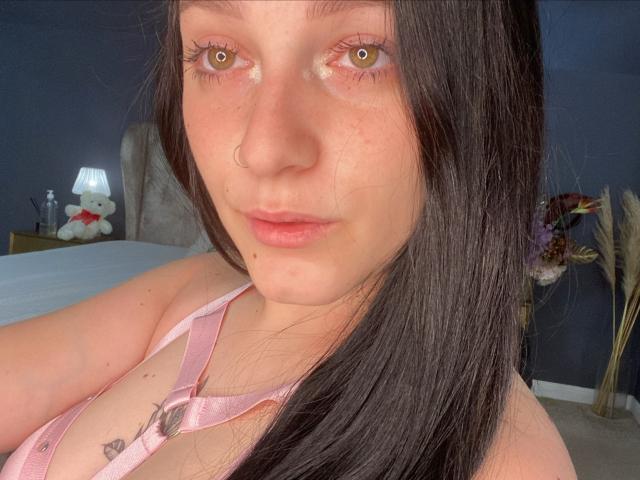 LeahBrooks with long black hair, pink halter top, nose ring, and tattoo on chest, looking at camera with pale skin, subtle freckles, and pinkish lips, in bedroom.