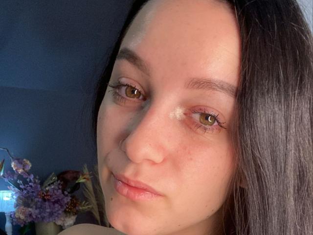 LeahBrooks has pale skin, brown eyes, long black hair, and a slight smile with subtle freckles on her nose. Her eyelashes are long and she has a small beauty mark.