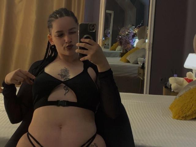 LeahBrooks with braided hair, wearing black lingerie with cutouts, showing her tits and belly, taking a mirror selfie in a dimly lit room, holding a phone.