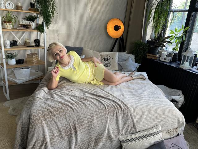 OliviaHorst with blonde hair, yellow dress, and big tits, laying on a bed with a grey patterned blanket, holding a straw, in a cozy, plant-filled bedroom.