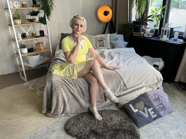 OliviaHorst, blonde, yellow dress, short sleeves, white socks, sitting on bed, legs crossed, ass visible, pensive, soft smile, cozy bedroom, plants, shelf, round rug, fluffy blanket.