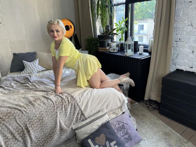 OliviaHorst, blonde, cute, yellow dress, kneeling on bed, white socks, smiling, ass up, bedroom, gray bedspread, black radiator, green plant, white brick wall, daytime, natural light.