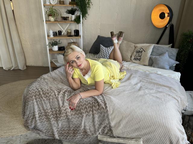 OliviaHorst, blonde, laying on bed in yellow dress, showing ass up, legs crossed, looking at camera, red nails, wooden floor, gray and beige blankets, large orange studio light.