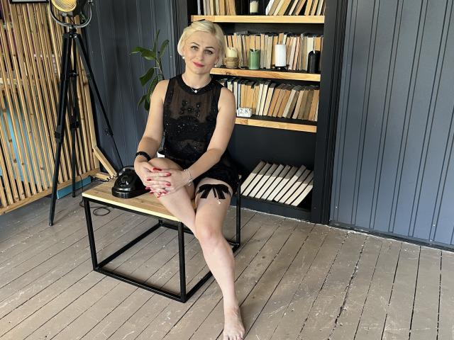 OliviaHorst with blonde hair, wearing black lace dress, sitting on wooden table, legs crossed, showing her bare-ass and black garter on right thigh, red nails, black phone.