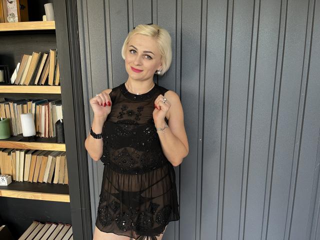 OliviaHorst with blonde hair, wearing a sheer black dress with floral lace, standing, hands up, red nails, smiling, gray wall, bookshelf with books on left.