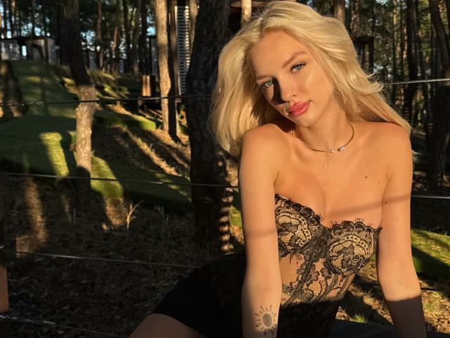 LexyGlam with blonde hair, blue eyes, and red lipstick, wearing a black lace strapless top, sitting in a forest, sunlight casting shadows, small tattoo on right arm.