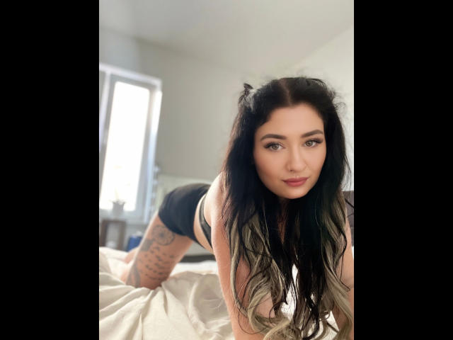Claudiliciousxx-hot on all fours, wearing black lingerie, with cat ears, long black-and-blonde hair, tattooed thigh, and a sultry smile, on a bed.