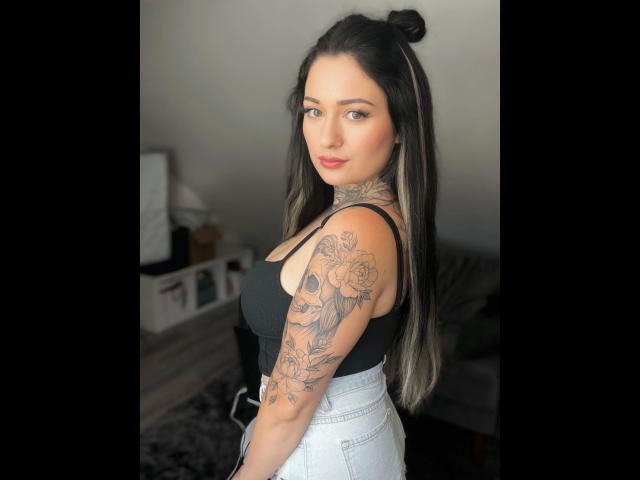 Claudiliciousxx-hot with long black hair in a bun, black tank top, white high-waisted jeans, tattooed arm, looking over shoulder, red lipstick, standing in a room.