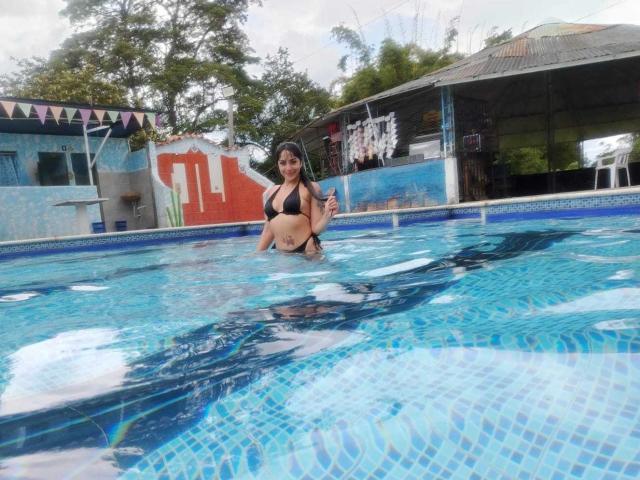 AnalisseJade in a blue-tiled pool, wearing a black bikini, has large boobs and a tattoo on her right thigh, holding a drink, with a roofed area in the background.
