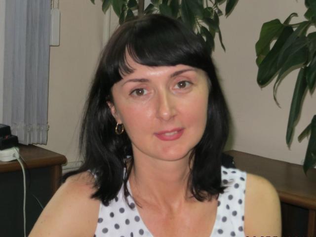 VivianGuchih has black shoulder-length hair, green eyes, and is wearing a white sleeveless top with black polka dots, with small gold earrings, and a slight smile.