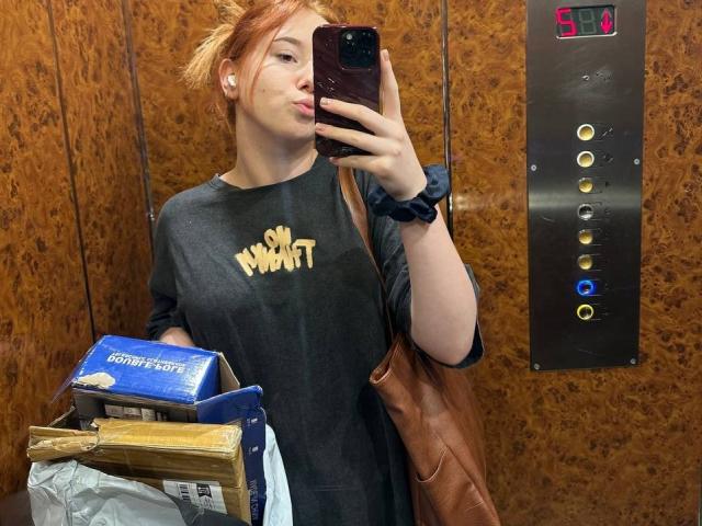 Barberry with red hair, pouty lips, holding phone, wearing black "So Wrong" shirt, brown bag, and blue scrunchie, standing in elevator, holding boxes, elevator buttons visible.
