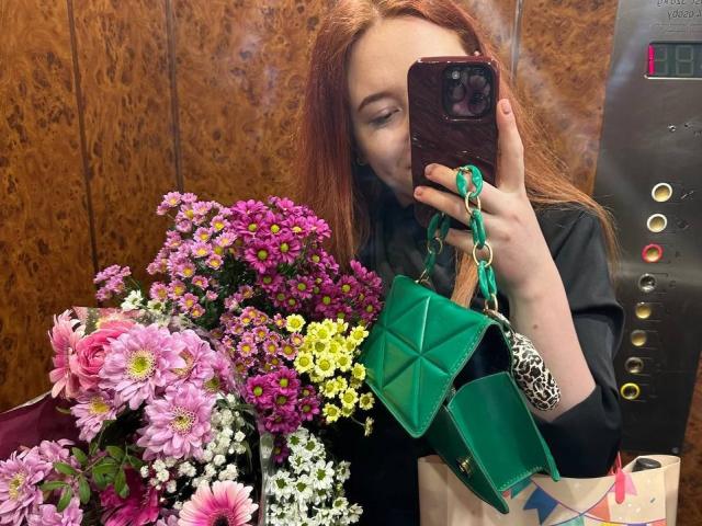 Barberry with red hair, black shirt, holding green purse, taking selfie in elevator, surrounded by pink, purple, yellow flowers, using phone with brown case, visible elevator buttons.