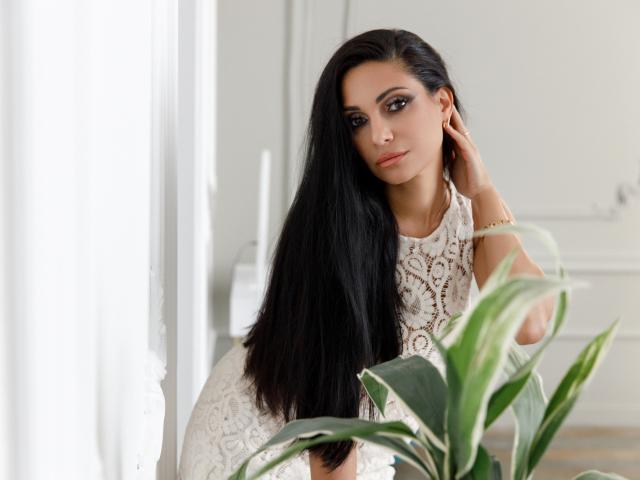 AllaYertlyi with long black hair, wearing a white lace dress, sitting near a green plant, looking at the camera with a slight smile, right hand touching her hair.