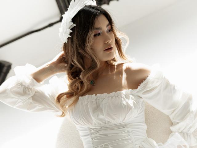 AnneTLuis with wavy blonde hair, white off-shoulder dress, puffed sleeves, floral headpiece, and necklace, looking down with soft lighting. Boobs are visible under the dress.