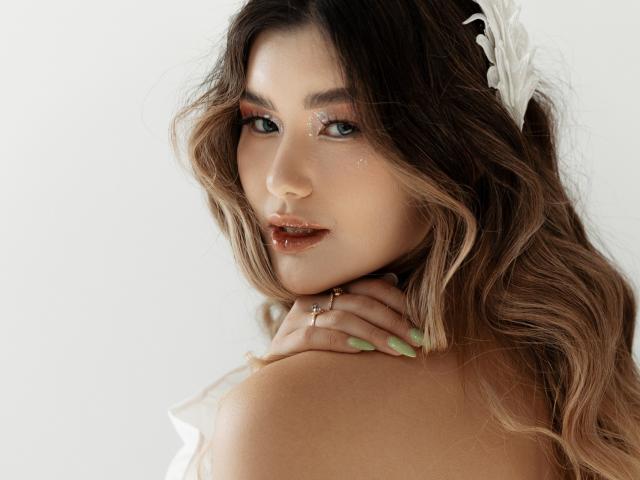 AnneTLuis has wavy brown hair, green nails, glossy brown lips, and a white flower headpiece. She's looking over her shoulder with a subtle smile, showing a ring on her neck.