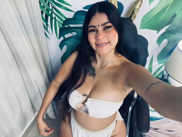 KeraniMarala mit langen, schwarzen Haaren, lächelnd, trägt einen weißen Bikini mit einem Ring in der Mitte, zeigt ihre Brüste, hat Tätowierungen an den Armen, sitzt auf einem Stuhl und macht ein Selfie..