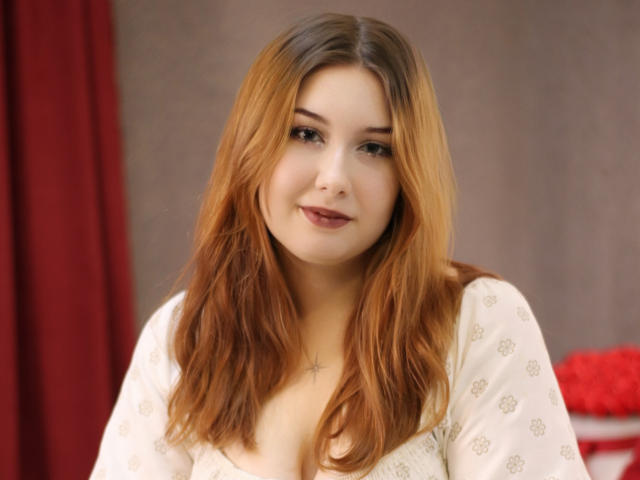TrishaKingsley has light brown, shoulder-length hair, a cross necklace, and a slight smile, wearing a white, patterned top, with visible, small, perky tits.