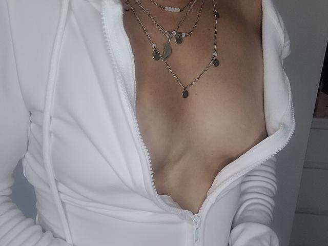 ElylaFrenchy's tits are exposed, showing cleavage, in a white zip-up hoodie, with a silver necklace featuring crescent moon and bead designs, against a gray background.