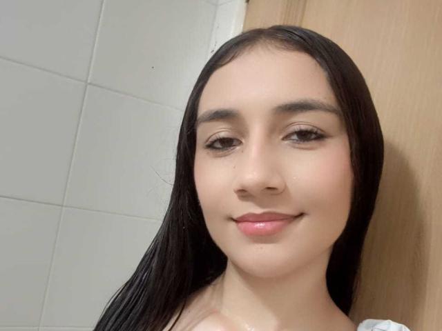 VaioletMiller has smooth, light brown skin, long black hair, and is smiling with pink lipstick. Her small tits are partially visible under a white shirt.
