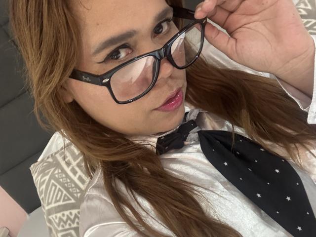 SophiaQueens, con lunghi capelli castani, che indossa occhiali, una camicia bianca e una cravatta nera con piccoli motivi bianchi, tiene gli occhiali con la mano destra, rossetto rosa..