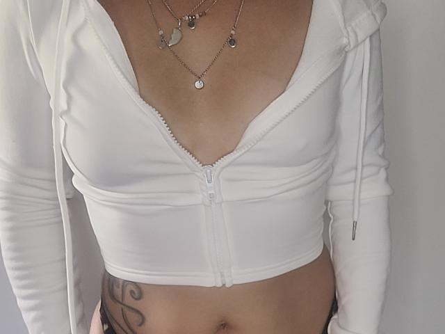 ElylaFrenchy wearing a white zip-up top that reveals her tits, with a necklace and tattoo on her right side. Her skin is light and her top is unzipped.