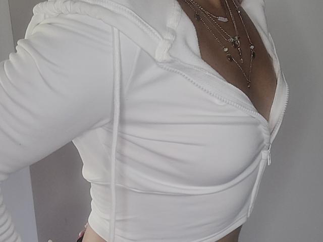 ElylaFrenchy's white zip-up top, showing her tits, with a silver necklace and her right arm raised. Her top is tight, highlighting her cleavage and midriff.