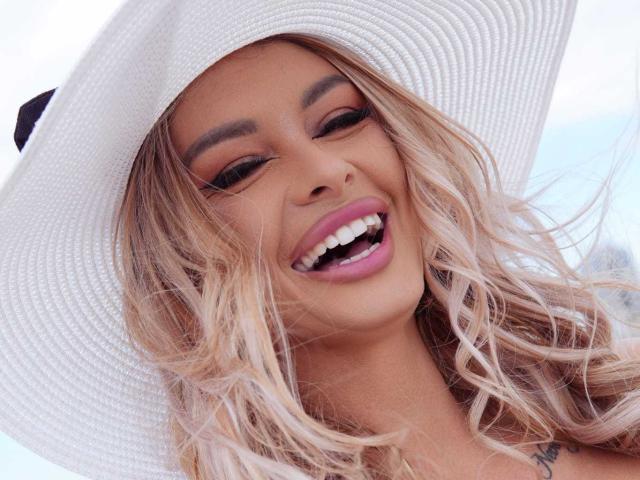 OneDiamond with blonde wavy hair, white sunhat, and pink lipstick, smiling with open mouth, showing white teeth. OneDiamond's eyes are partially closed. OneDiamond's tits are partially visible.